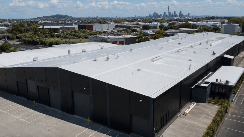 A large industrial warehouse in Auckland, New Zealand, with a freshly restored white metal roof coating system, dark exterior walls, surrounding commercial buildings, and the city skyline in the distance.